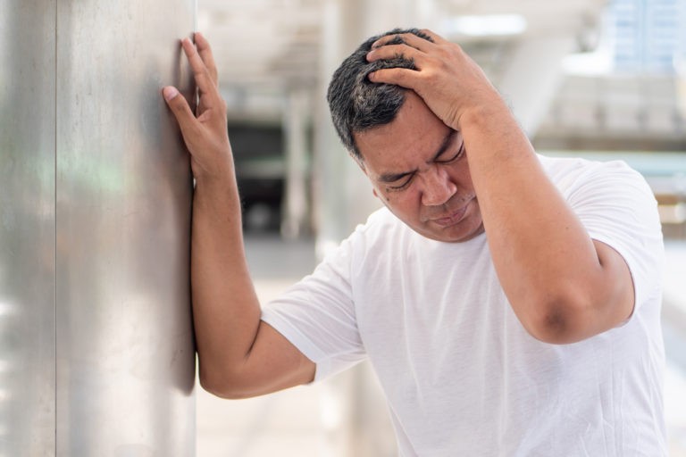 senior asian man leaning on wall with head covered by left hand due to vertigo