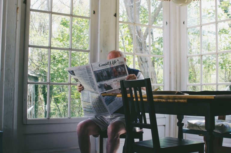 man over 65 sitting in kitchen with sprawling windows reading Good Life newspaper